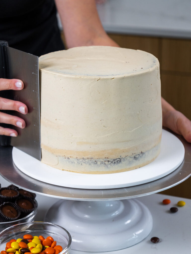 image of peanut butter frosting being smoothed onto a reese's chocolate peanut butter cake with a bench scraper