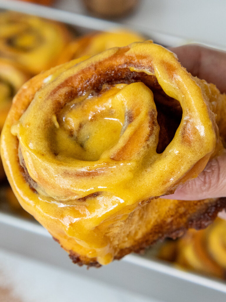 image of a pumpkin pie cinnamon roll being held up to show its pumpkin cream cheese icing