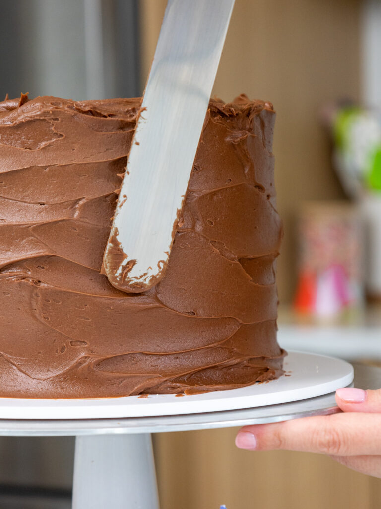 image of chocolate buttercream being spread around a yellow layer cake with a large offset spatula