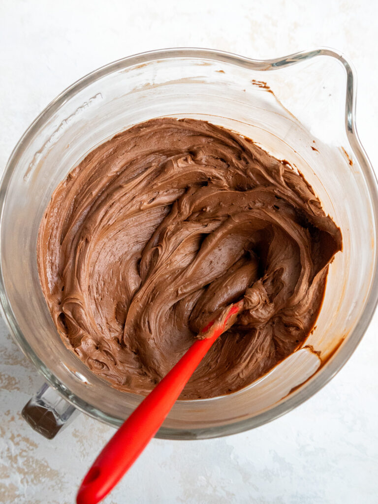 image of chocolate buttercream that's been stirred in a large glass mixing bowl with a red spatula to make it smooth
