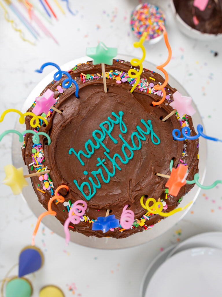 image of a yellow cake frosted with chocolate buttercream that's been decorated with fun candles and has had happy birthday written on top of it with frosting