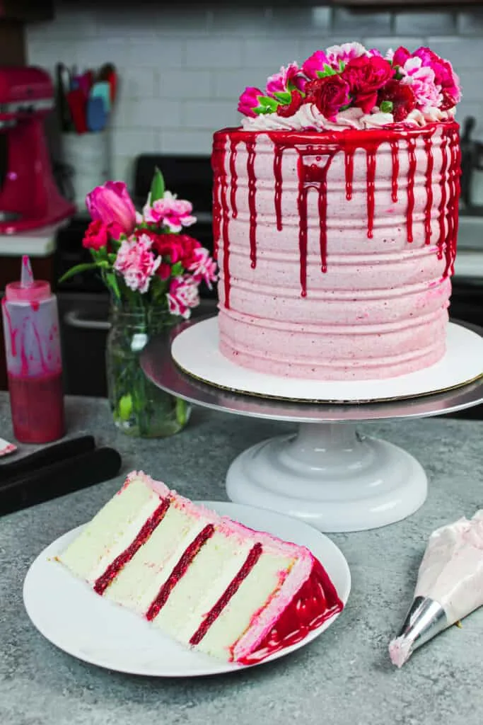 image of raspberry white chocolate cake decorated with fresh raspberries, flowers, and a white chocolate drip with slice in front