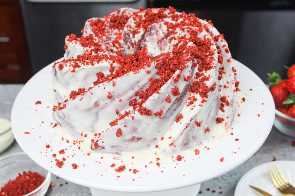 image of beautiful red velvet bundt cake, completely covered in cream cheese glaze and decorated with red velvet crumbs on top