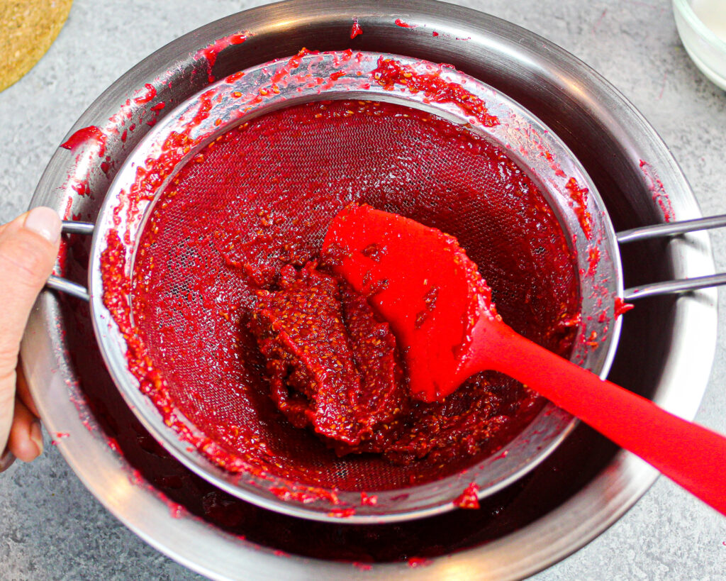 image of raspberry cake filling being pushed through a fine mesh sieve to remove the seeds