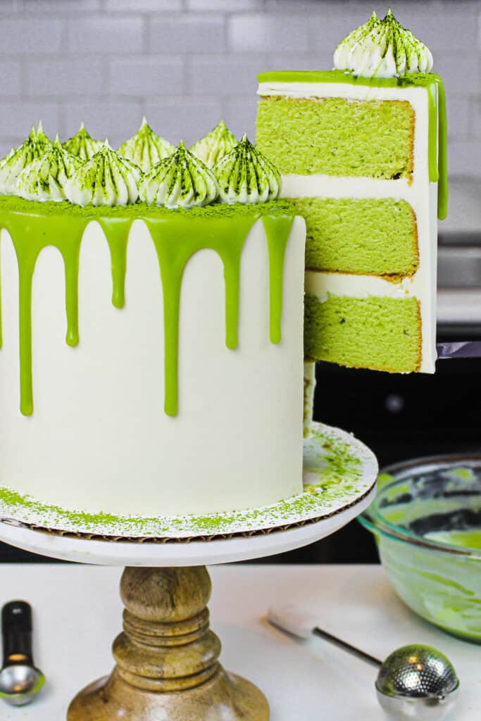 image of matcha cake slice on a plate with pretty matcha drip cake in background