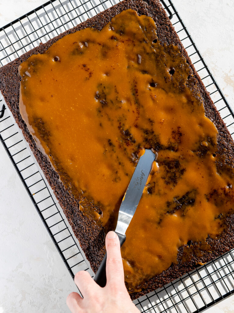 image of warm caramel being spread over a chocolate sheet cake to make a chocolate caramel poke cake