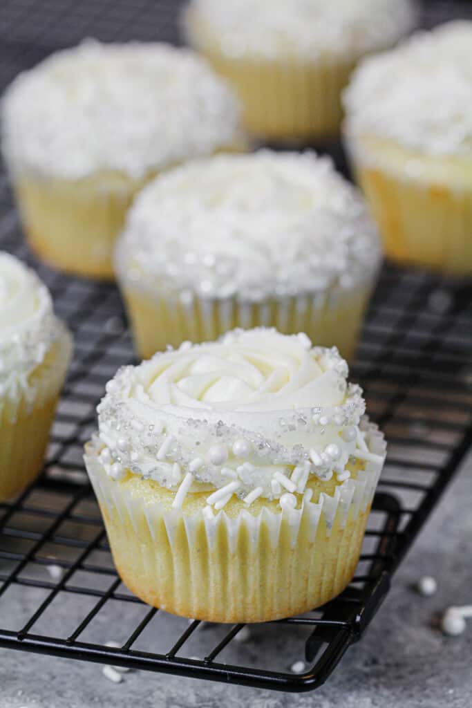 image of almond cupcakes on wire rack frosted and decorated with a white sprinkle blend