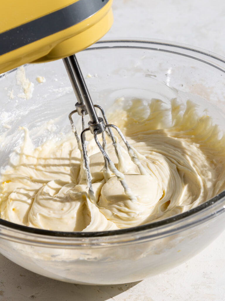 image of lemon cream cheese frosting being mixed in a glass bowl with a yellow kitchenaid hand mixer