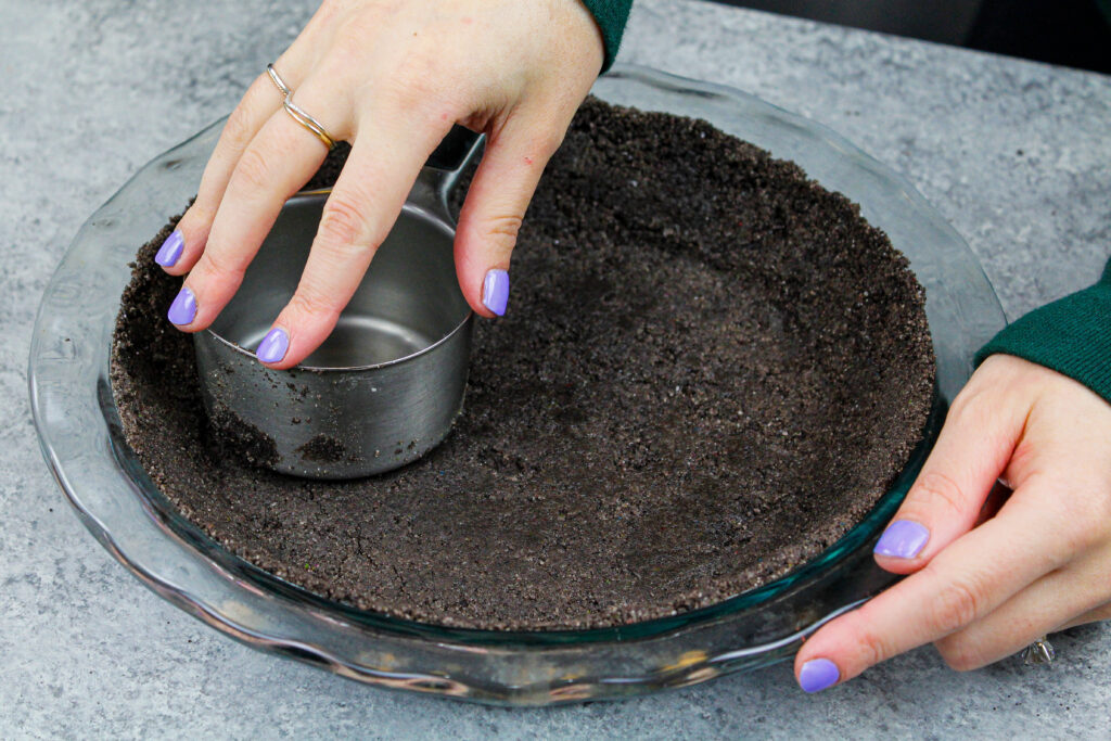 image of an oreo pie crust being made and pressed into a pie pan using a metal measuring cup