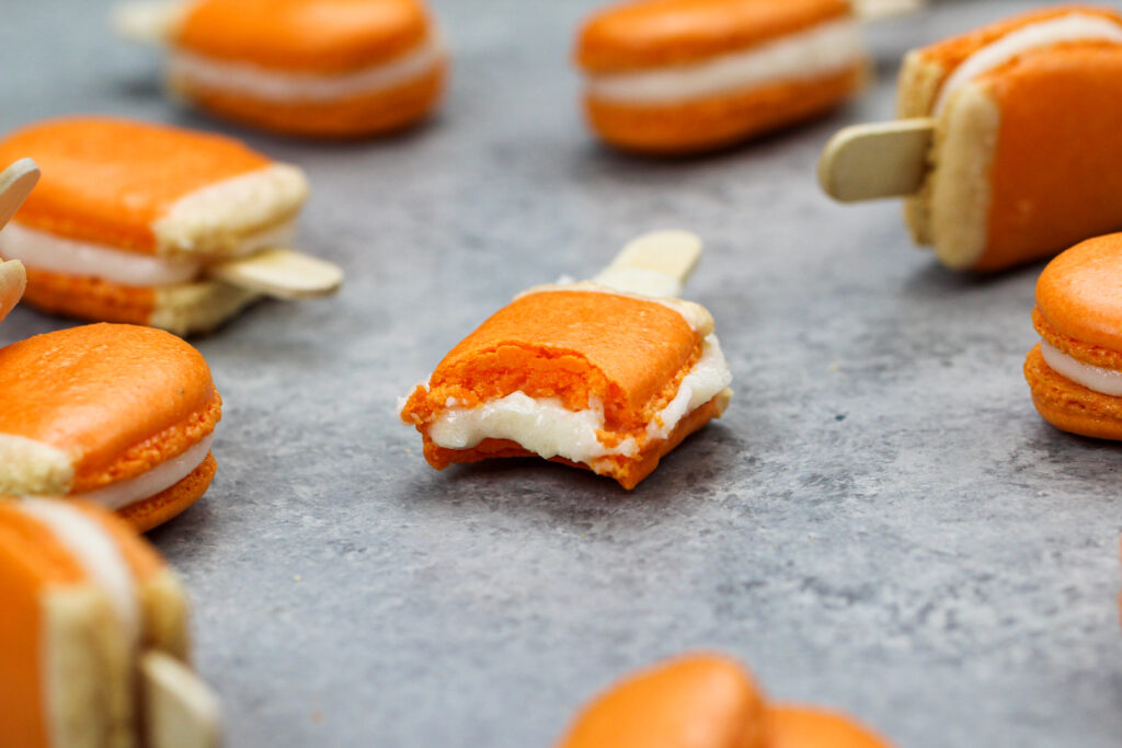 image of mini orange creamsicle macarons filled with orange creamsicle buttercream that have been bitten into to show the filling and full shells