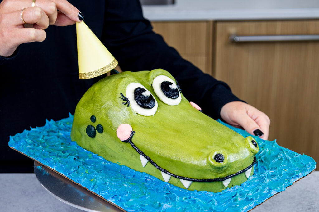 image of a party hat being added to a crocodile cake by chelsey white of chelsweets as part of her animal cake series