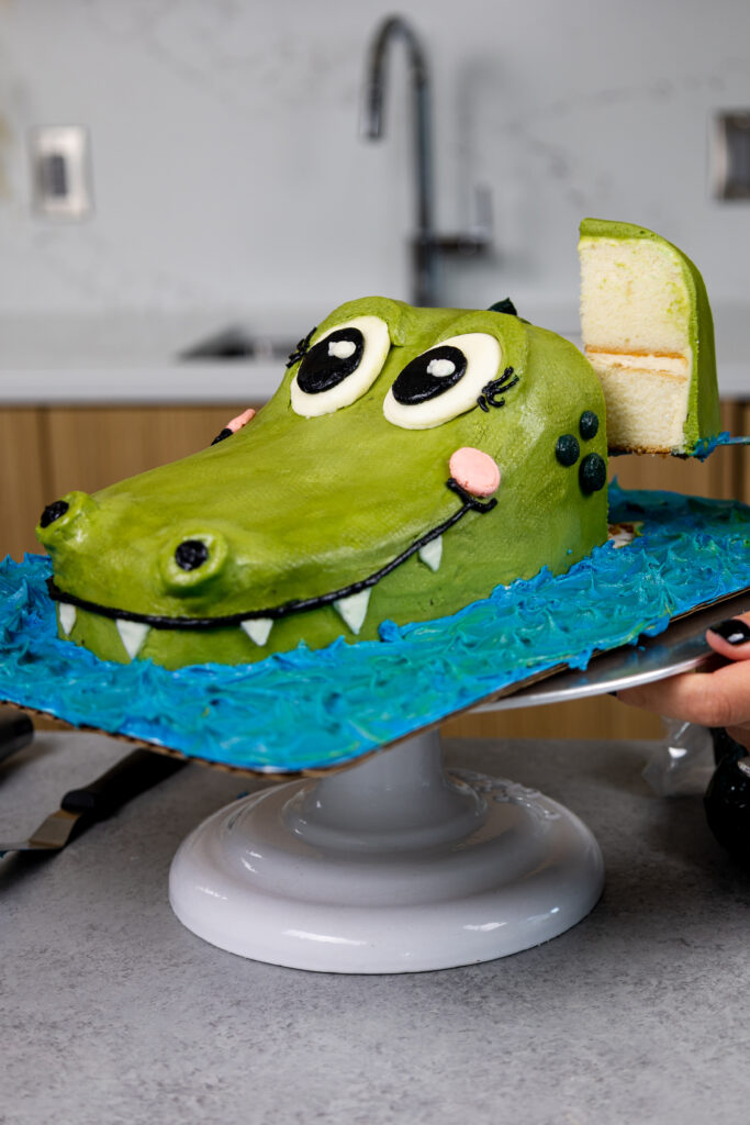 image of a slice of cake being cut from a cute crocodile cake made with buttercream by chelsweets
