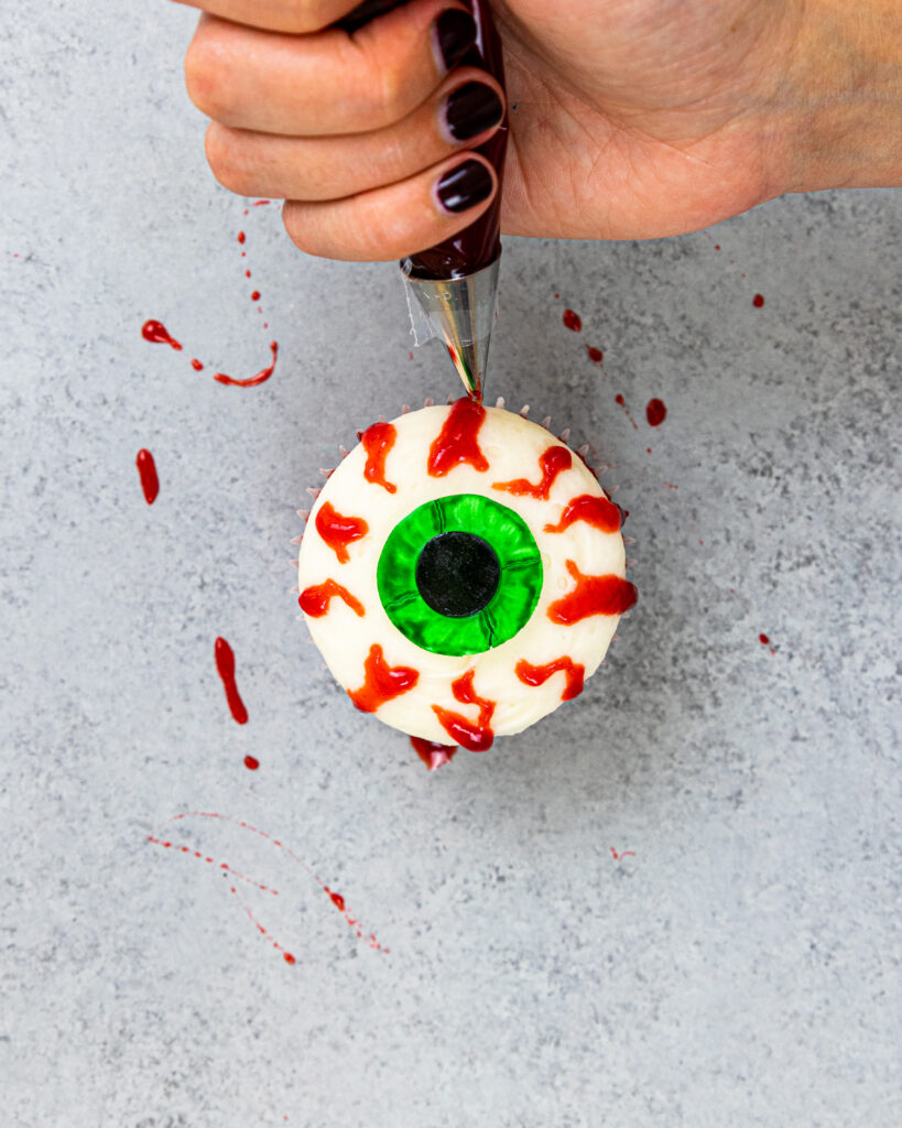 image of edible blood made with raspberry jam being used to pipe veins on the side of an eyeball cupcake