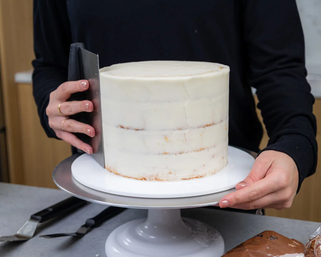 image of a vanilla cake being crumb coated with buttercream frosting using a bench scraper