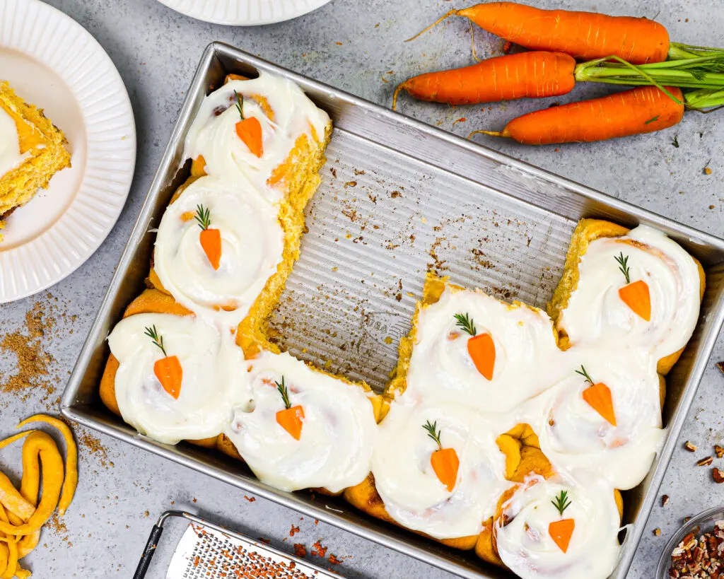 image of carrot cake cinnamon rolls in a pan that have been frosted with cream cheese frosting and decorated with cute little carrots