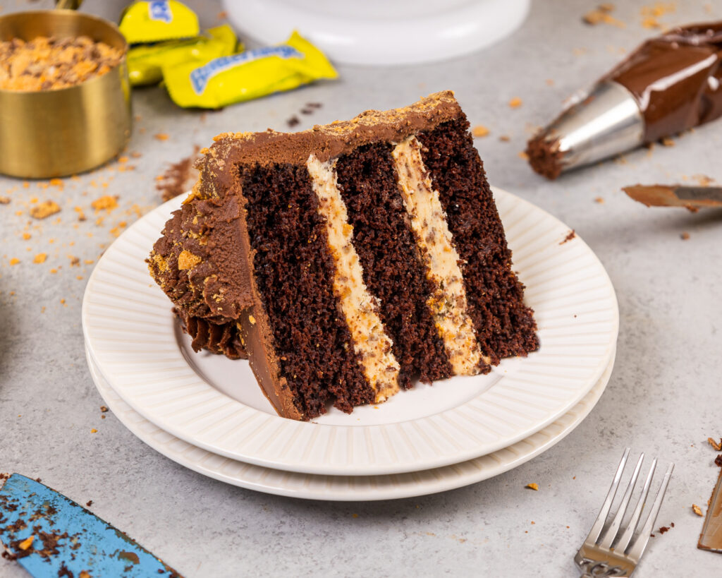 image of a slice of butterfinger cake on a plate that's made with tender chocolate cake layers, crunchy peanut butter butterfinger filling and frosted with chocolate buttercream