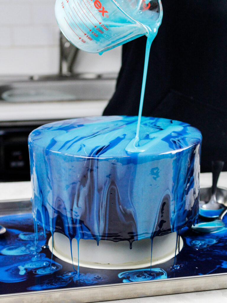 image of blue mirror glaze being poured over a chilled buttercream cake