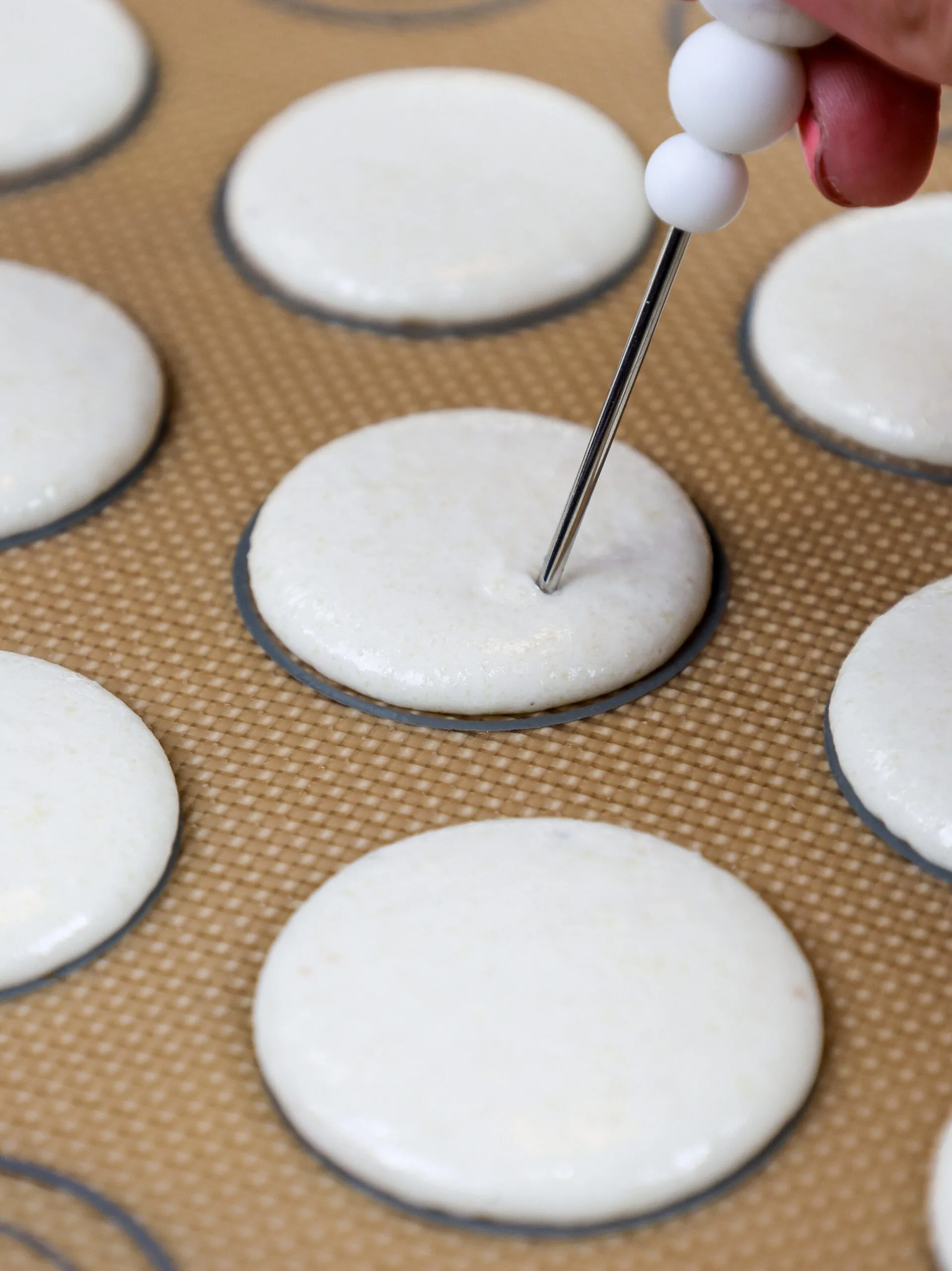 image of white vanilla macaron shells that been been banged against the counter to release air bubbles and are having any air bubbles that have surfaced by popped with a scribe