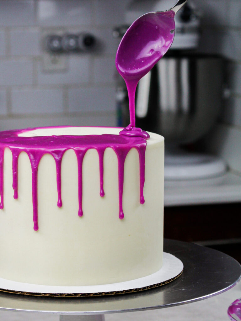image of white chocolate ganache drips being added to a chilled buttercream cake to make a purple drip cake