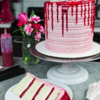 image of raspberry white chocolate cake decorated with fresh raspberries, flowers, and a white chocolate drip with slice in front