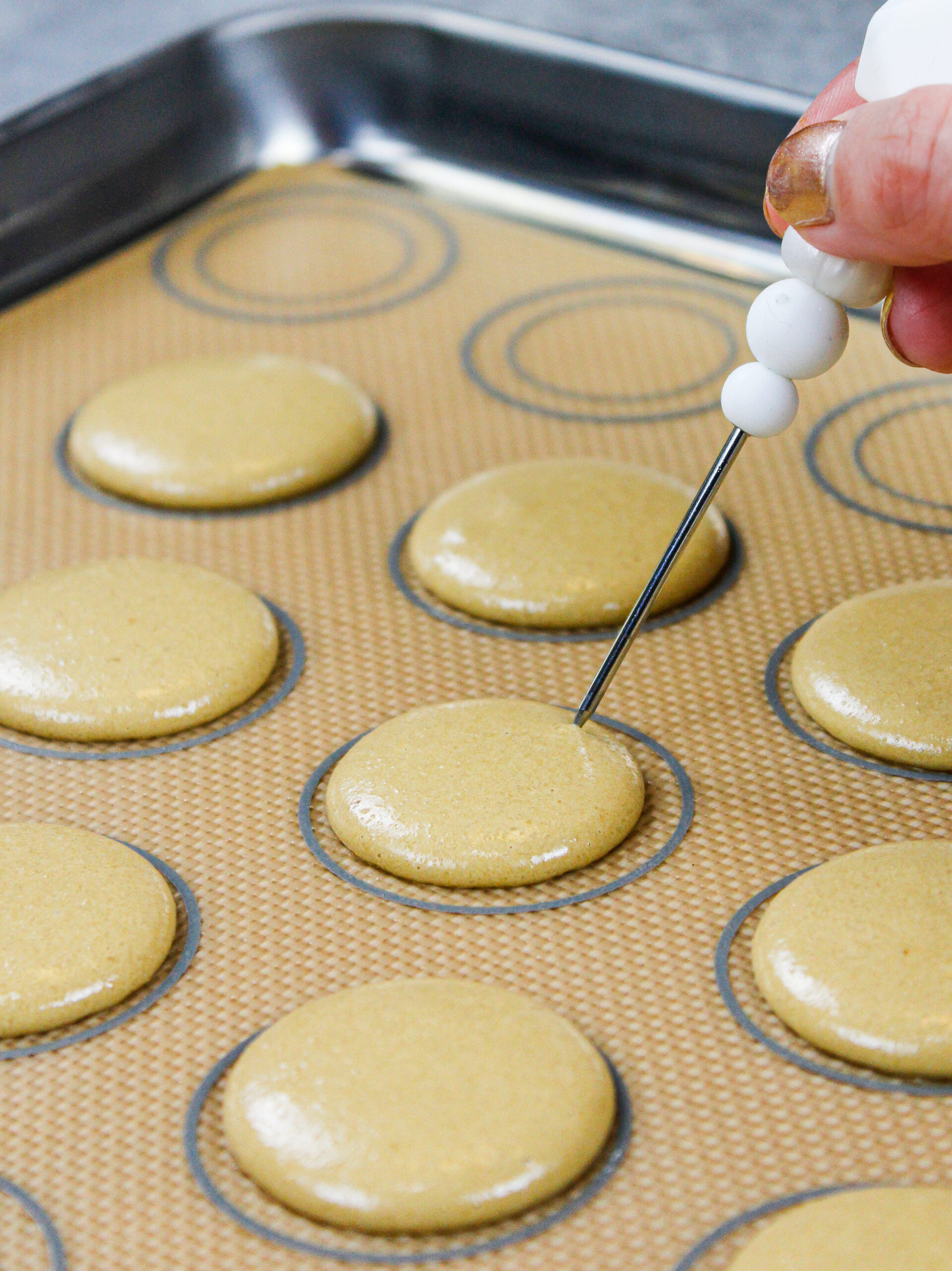 image of coffee macaron shells that have been piped and banged on a counter to release any air bubbles, which are being popped with a scribe