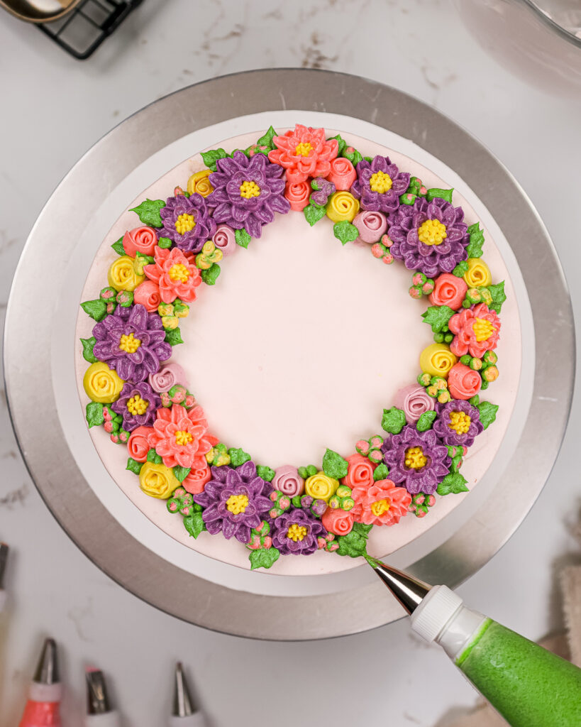 image of buttercream leaves being piped onto a buttercream flower cake to fill in any gaps between the flowers