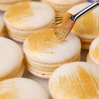 image of a white chocolate macaron that's been filled with white chocolate ganache and decorated with edible gold paint streaks
