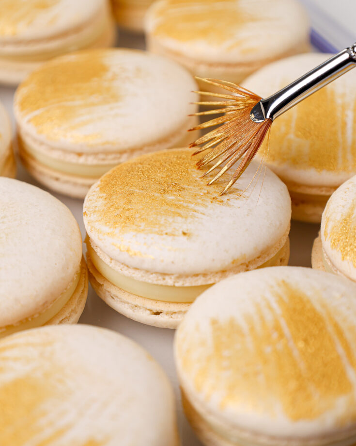 image of a white chocolate macaron that's been filled with white chocolate ganache and decorated with edible gold paint streaks