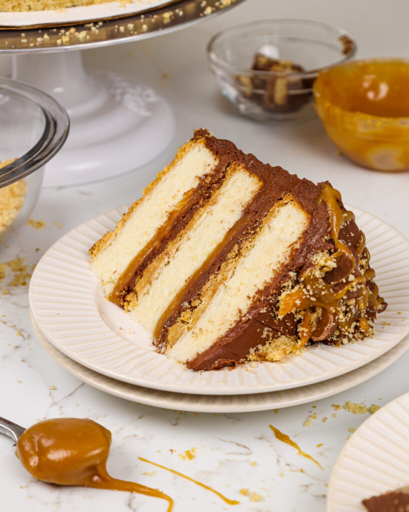 image of a slice of a Twix cake on a plate, showing its shortbread crust, vanilla cake layers, thick caramel filling, and chocolate buttercream