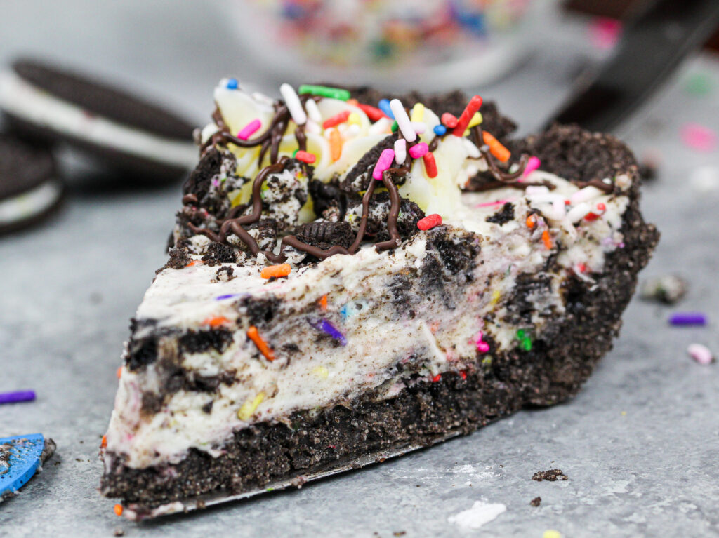 image of a slice of oreo cream pie decorated with sprinkles and a chocolate drizzle that's shared as part of a no bake recipe round up