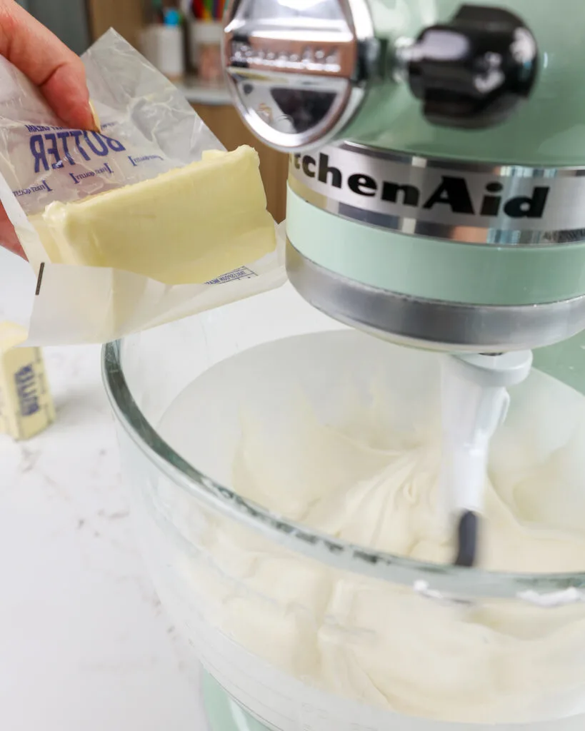 image of a stick of room temperature butter being added into meringue to make a perfectly sweet frosting