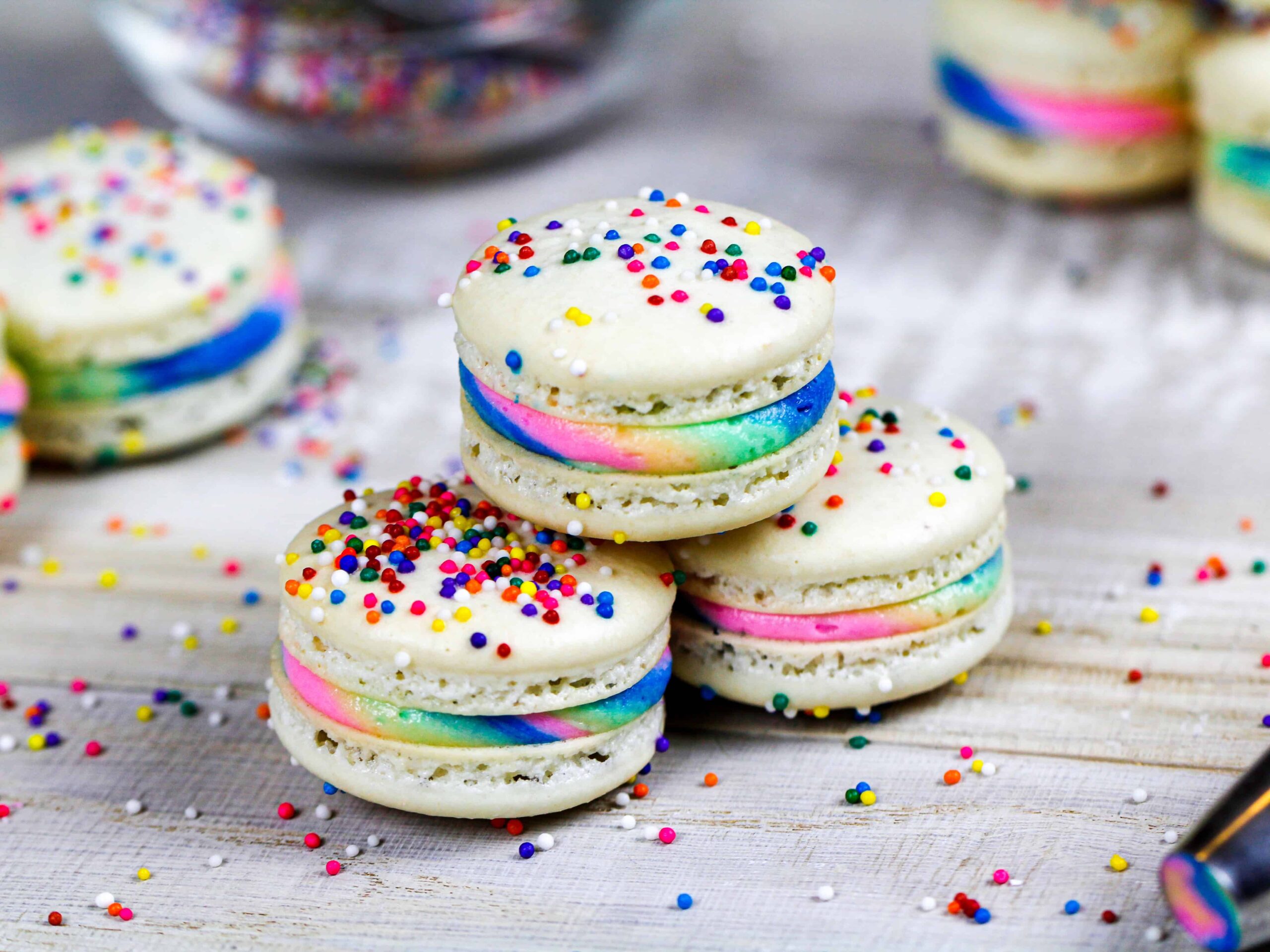 image of three rainbow macarons stacked on top of each other to show off their sprinkle topping and rainbow buttercream