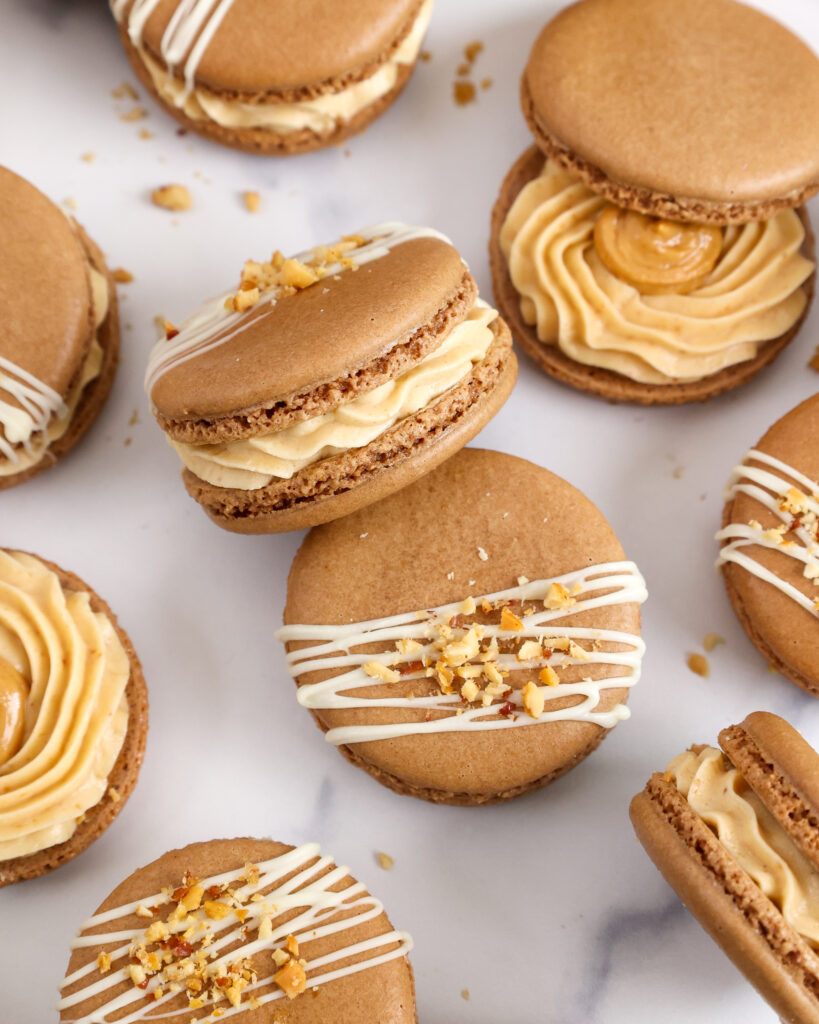 image of peanut butter macarons filled with honey roasted peanut butter buttercream and topped with chopped peanuts and a white chocolate drizzle