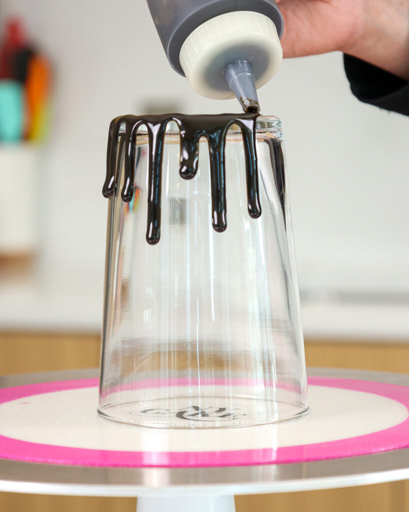 image of black drips being added around a glass to test the consistency of the ganache and make sure it's good to go before adding it to a black drip cake