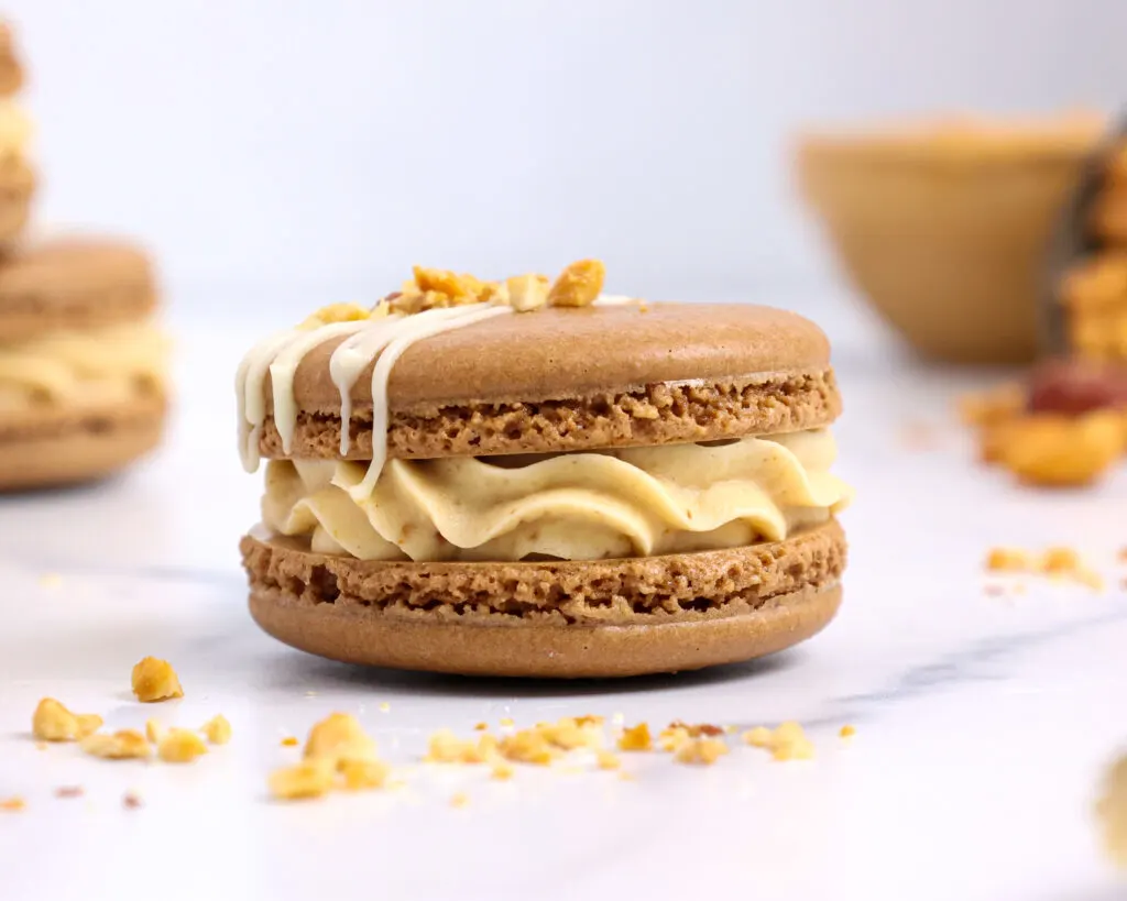 image of peanut butter macarons filled with honey roasted peanut butter buttercream and topped with chopped peanuts and a white chocolate drizzle