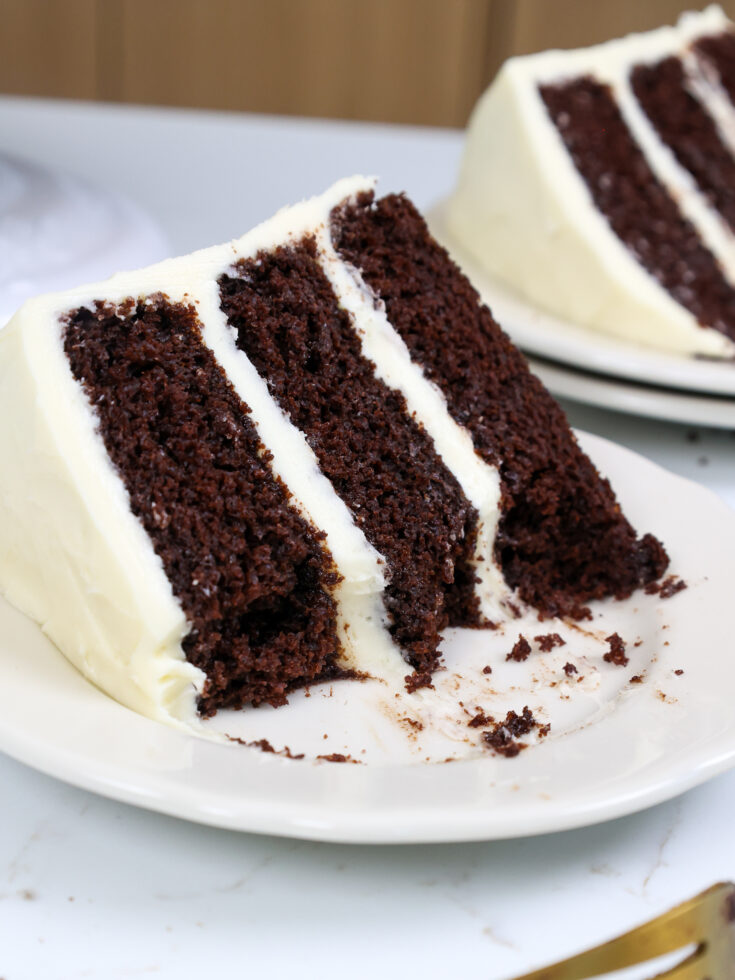 image of a slice of chocolate cake frosted with cream cheese frosting on a plate that's been cut into to show how moist it is