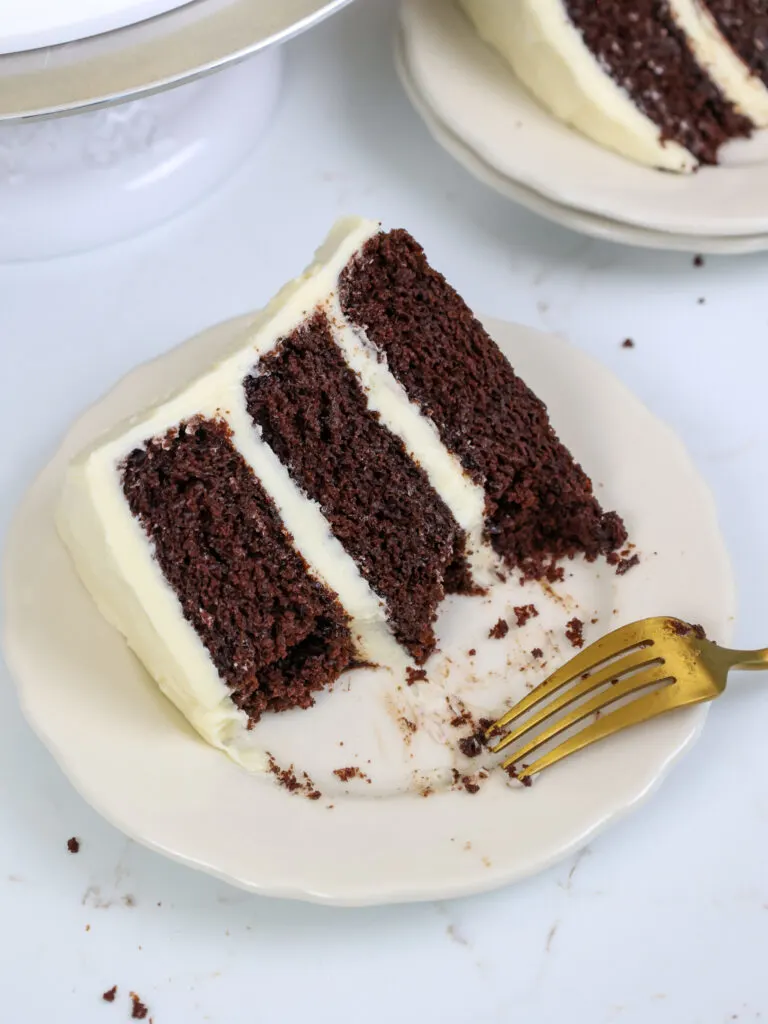 image of a slice of chocolate cake frosted with cream cheese frosting on a plate that's been cut into to show how moist it is