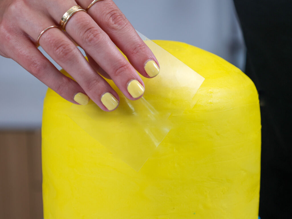 image of a cake being covered in thick layer of yellow buttercream and smoothed with an acetate sheet
