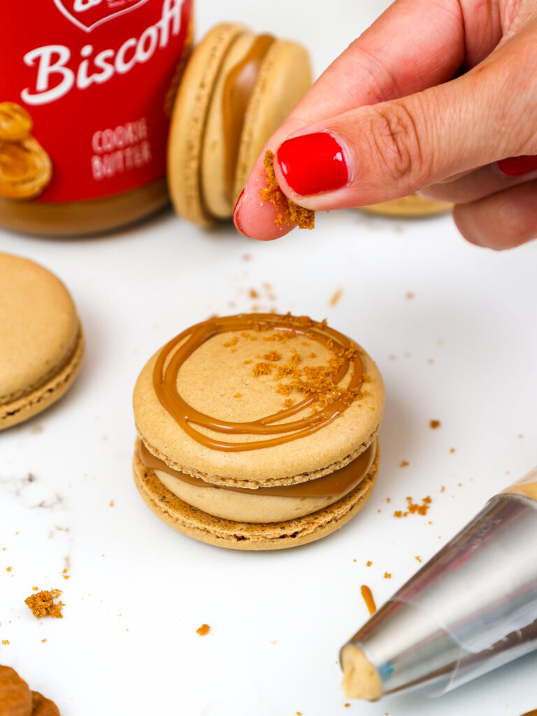 image of biscoff cookie crumbs being sprinkled over the top of a biscoff cookie butter macaron