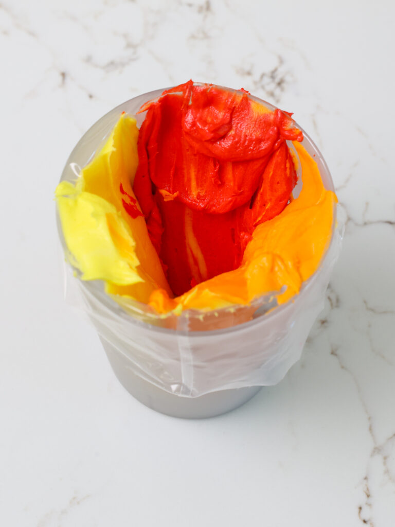 image of red, orange, and yellow buttercream in a frosting bag that will be used to pipe tri-colored swirls on a cake to look like flames