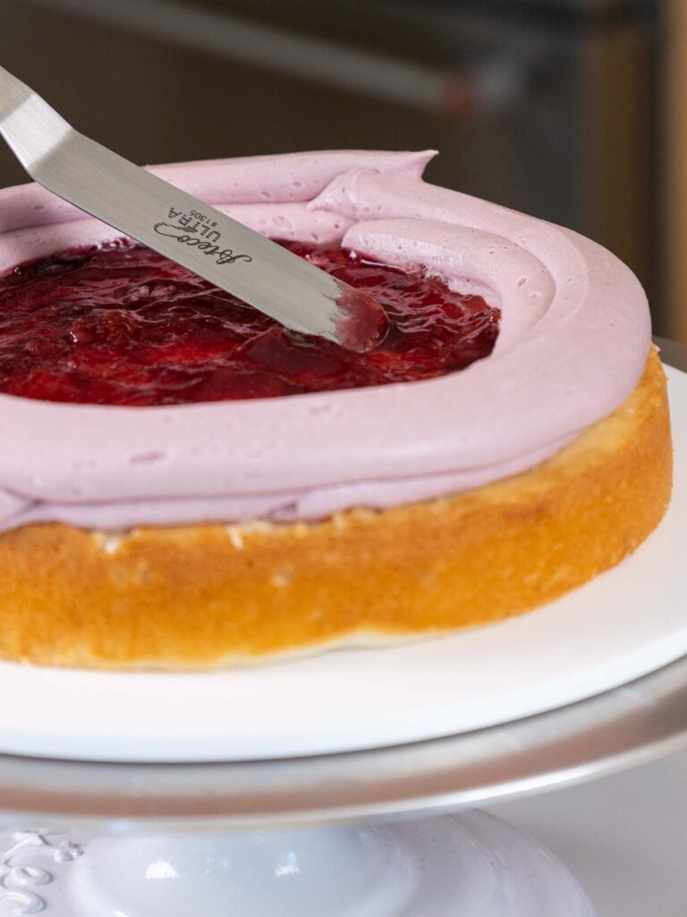 image of blackberry jam being smoothed onto a vanilla cake layers inside a ring of buttercream frosting
