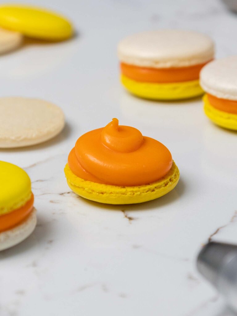 image of a yellow macaron shell being filled with candy corn ganache filling