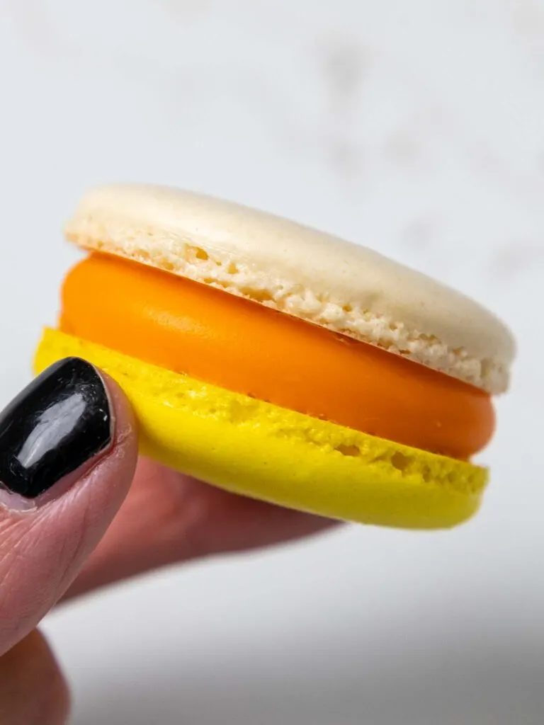 image of a candy corn macaron that have been filled with candy corn ganache that's being held up in the air