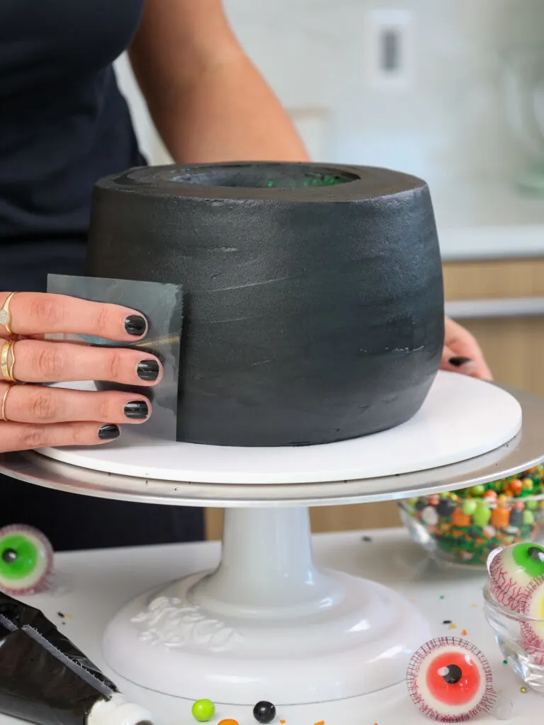 image of black buttercream being smoothed onto a cauldron cake with an acetate sheet