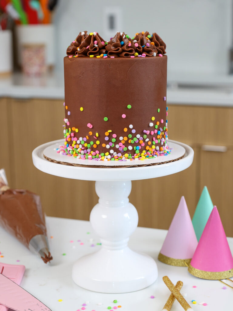 image of a small, 4-inch chocolate cake that's been frosted with chocolate buttercream that has been placed on a cute little white cake stand with party decorations around it