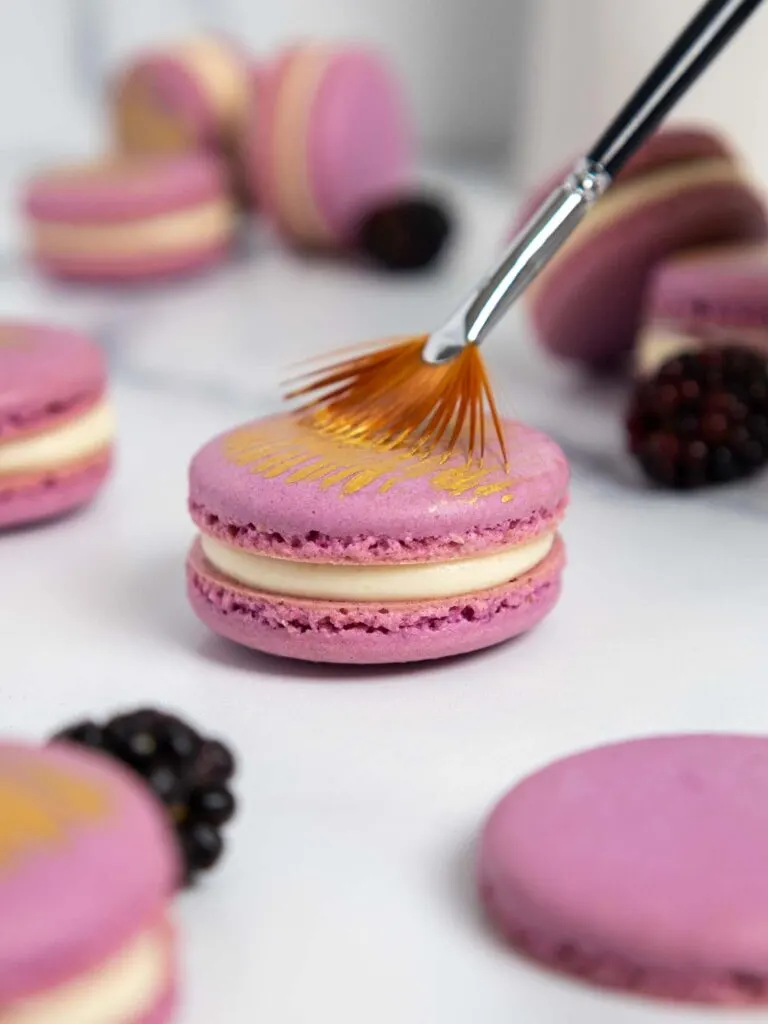 image of a blackberry macaron being decorated with a brush of edible gold paint