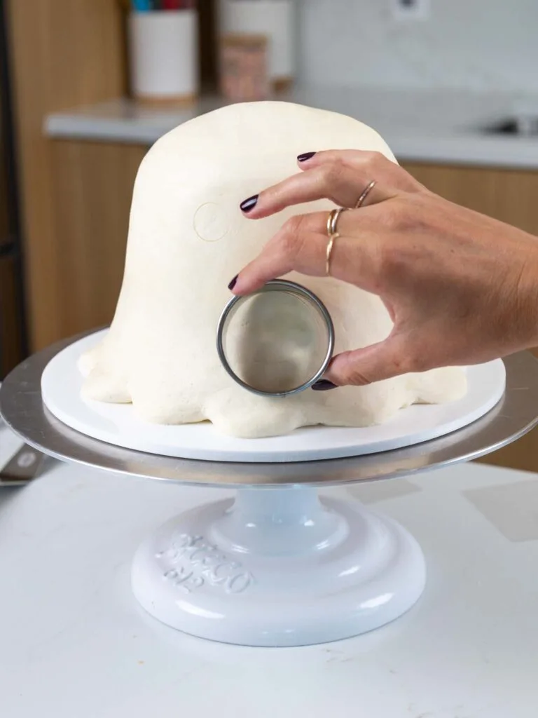 image of a circle cutter being gently pressed into a ghost shaped cake to outline where to pipe the ghost's face and pumpkin that it will hold