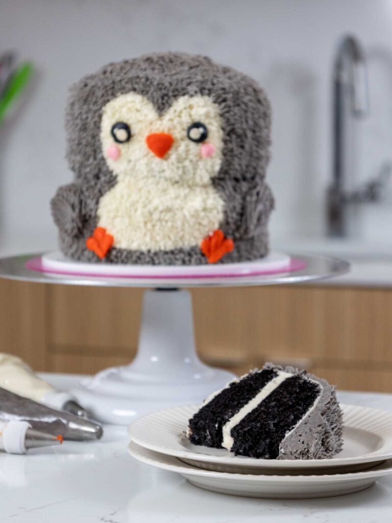 image of a slice of dark chocolate cake on a plate that's been cut from a cute buttercream penguin cake