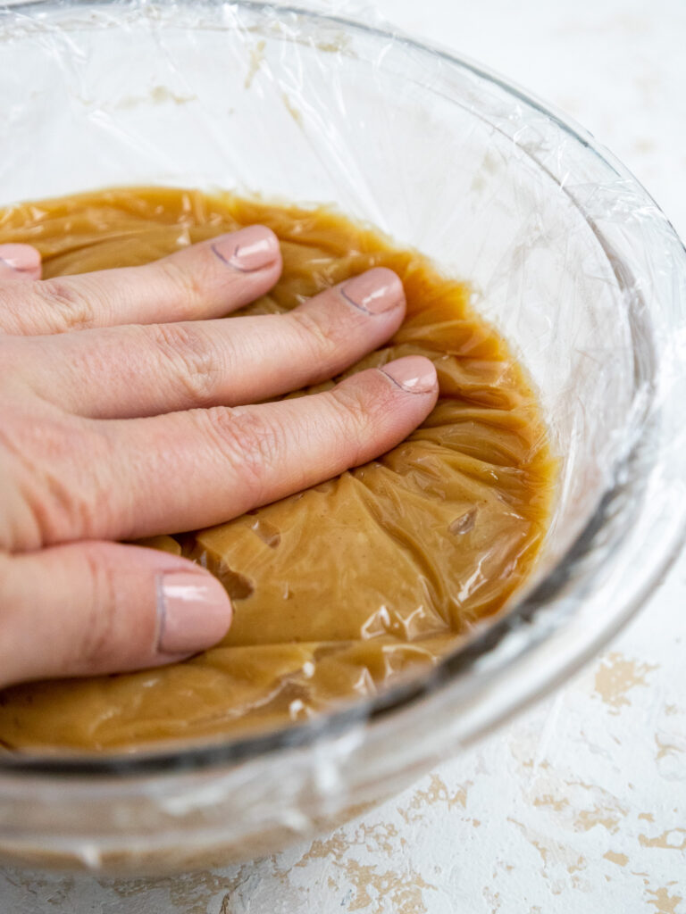 image of plastic wrap being pressed flush against peanut butter ganache to prevent it from forming a skin as it cools