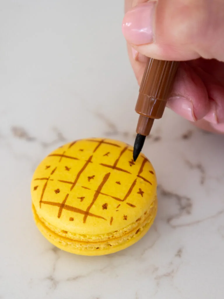 using a brown edible marker to draw criss crossed lines on a yellow macaron shell to make it look like a pineapple